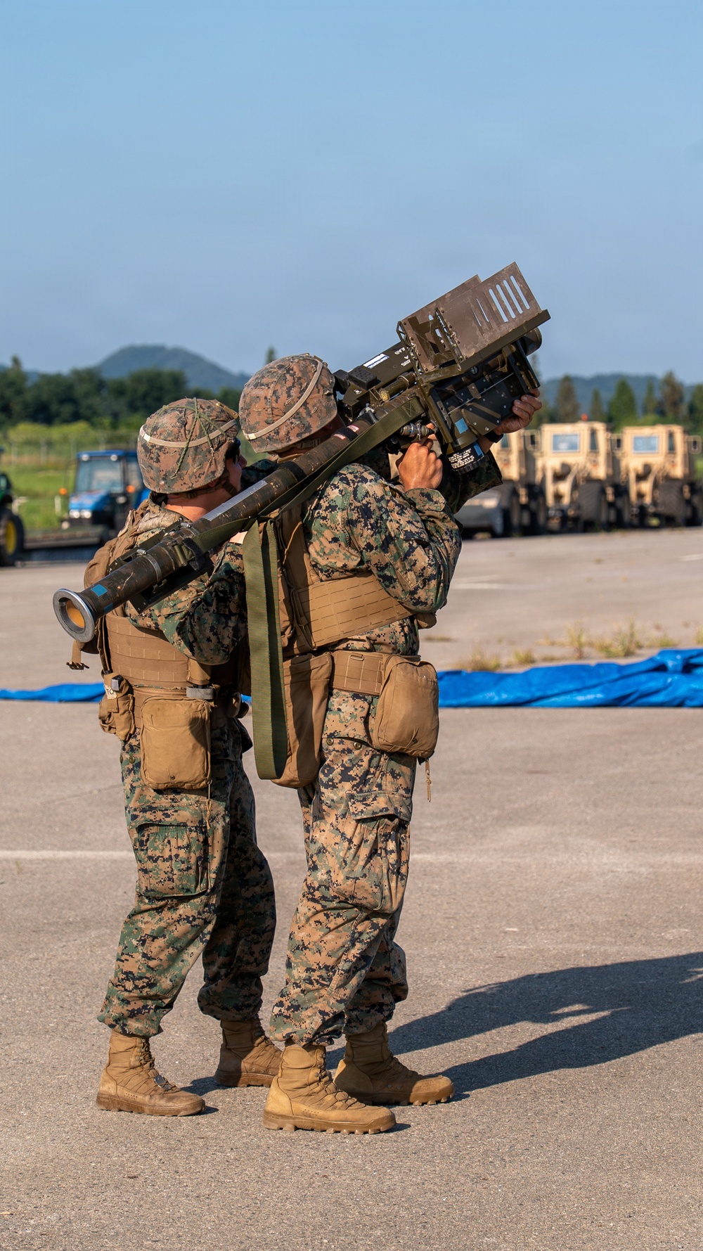 Two military personnel in MARPAT (Marine pattern) camouflage uniforms are operating what appears to be a man-portable air-defense system (MANPADS), likely a FIM-92 Stinger missile launcher. They are standing on a paved surface, possibly an airfield or military installation, with military vehicles visible in the background. The image appears to document a training exercise or demonstration.