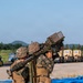 Two military personnel in MARPAT (Marine pattern) camouflage uniforms are operating what appears to be a man-portable air-defense system (MANPADS), likely a FIM-92 Stinger missile launcher. They are standing on a paved surface, possibly an airfield or military installation, with military vehicles visible in the background. The image appears to document a training exercise or demonstration.