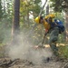 A wildland firefighter in full protective gear is working to extinguish a smoldering ground fire in a forested area. The individual is using a hand tool, likely a Pulaski or similar firefighting implement, to dig into and break apart burning debris on the forest floor. Smoke is rising from the disturbed soil and charred material, indicating active mop-up operations.