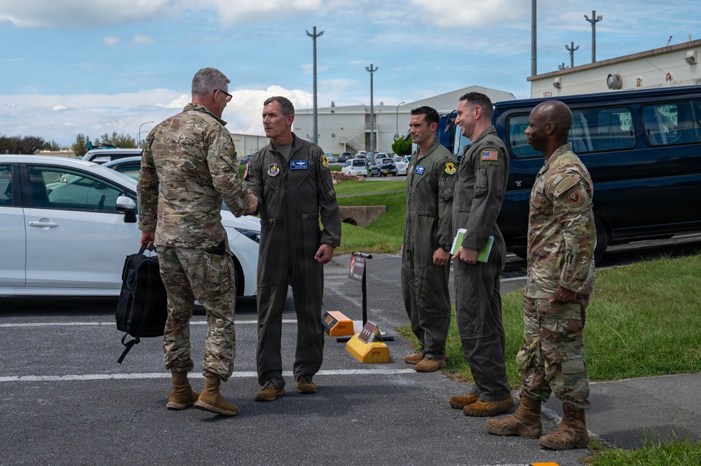 A group of five U.S. military personnel are gathered in a parking lot at what appears to be a military air base. Two individuals in camouflage (OCP) uniforms are interacting with three personnel wearing olive drab flight suits. A handshake is taking place between the leftmost OCP-uniformed individual and the nearest flight-suited individual, suggesting a greeting or formal meeting.