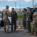 A group of five U.S. military personnel are gathered in a parking lot at what appears to be a military air base. Two individuals in camouflage (OCP) uniforms are interacting with three personnel wearing olive drab flight suits. A handshake is taking place between the leftmost OCP-uniformed individual and the nearest flight-suited individual, suggesting a greeting or formal meeting.