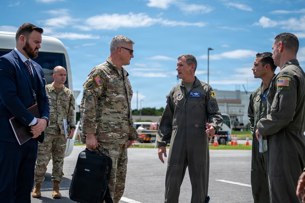 A group of U.S. military personnel and a civilian are gathered on what appears to be a military airfield tarmac. Two senior officers — one in Army OCP camouflage uniform and one in an Air Force flight suit — appear to be engaged in a formal greeting or briefing. The setting suggests an official arrival or reception ceremony at a military installation.