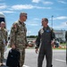A group of U.S. military personnel and a civilian are gathered on what appears to be a military airfield tarmac. Two senior officers — one in Army OCP camouflage uniform and one in an Air Force flight suit — appear to be engaged in a formal greeting or briefing. The setting suggests an official arrival or reception ceremony at a military installation.