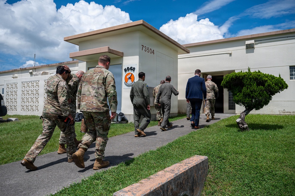 A group of military personnel and at least one civilian in a business suit are walking toward the entrance of Building 73504, which displays a 'HOGS' unit patch/emblem on its facade. The individuals are wearing a mix of camouflage uniforms (OCP pattern), flight suits, and civilian attire, suggesting a multi-service or official visit. The setting appears to be a military installation, likely an air base given the flight suits and unit insignia.