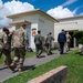 A group of military personnel and at least one civilian in a business suit are walking toward the entrance of Building 73504, which displays a 'HOGS' unit patch/emblem on its facade. The individuals are wearing a mix of camouflage uniforms (OCP pattern), flight suits, and civilian attire, suggesting a multi-service or official visit. The setting appears to be a military installation, likely an air base given the flight suits and unit insignia.