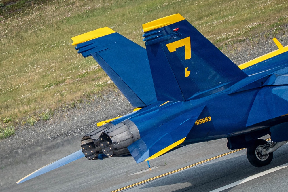 A close-up aerial photograph of a U.S. Navy Blue Angels F/A-18 Hornet jet aircraft numbered '7' on a runway. The aircraft displays the iconic blue and gold color scheme with Bureau Number 165663 visible on the fuselage. The jet appears to be in motion, with engine exhaust and heat distortion visible from the rear nozzle.
