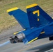 A close-up aerial photograph of a U.S. Navy Blue Angels F/A-18 Hornet jet aircraft numbered '7' on a runway. The aircraft displays the iconic blue and gold color scheme with Bureau Number 165663 visible on the fuselage. The jet appears to be in motion, with engine exhaust and heat distortion visible from the rear nozzle.