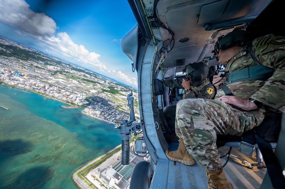 Two military personnel in camouflage uniforms are seated inside an open-door military helicopter flying over a coastal city with turquoise waters visible below. The image is taken from inside the aircraft looking outward, capturing both the crew and a sweeping aerial view of what appears to be an urban coastal area. A mounted weapon system is visible on the helicopter's door frame.