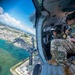 Two military personnel in camouflage uniforms are seated inside an open-door military helicopter flying over a coastal city with turquoise waters visible below. The image is taken from inside the aircraft looking outward, capturing both the crew and a sweeping aerial view of what appears to be an urban coastal area. A mounted weapon system is visible on the helicopter's door frame.