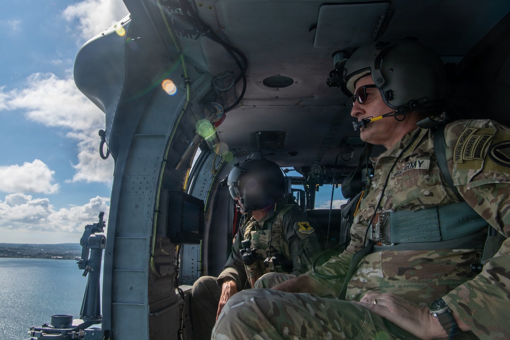 Two military personnel are seated inside a helicopter in flight, observing terrain below over a coastal area. The individual on the right wears U.S. Army insignia with what appears to be Special Forces and Ranger tabs, along with a communications headset. The image captures an aerial reconnaissance or survey mission over a body of water near a populated coastline.