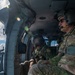 Two military personnel are seated inside a helicopter in flight, observing terrain below over a coastal area. The individual on the right wears U.S. Army insignia with what appears to be Special Forces and Ranger tabs, along with a communications headset. The image captures an aerial reconnaissance or survey mission over a body of water near a populated coastline.