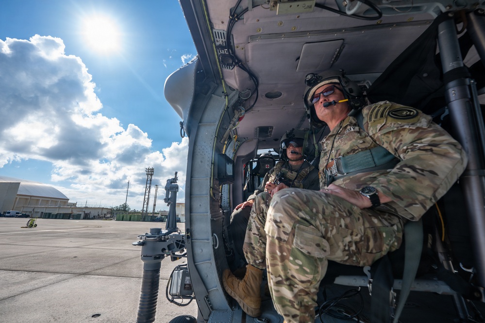Two military personnel in MultiCam/OCP camouflage uniforms are seated inside what appears to be a UH-60 Black Hawk helicopter on a tarmac. The soldier in the foreground wears a flight helmet with communications headset and sunglasses, while a second crew member is visible in the cockpit area. The aircraft appears to be on the ground at a military airfield with hangars and support structures visible in the background.