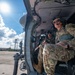 Two military personnel in MultiCam/OCP camouflage uniforms are seated inside what appears to be a UH-60 Black Hawk helicopter on a tarmac. The soldier in the foreground wears a flight helmet with communications headset and sunglasses, while a second crew member is visible in the cockpit area. The aircraft appears to be on the ground at a military airfield with hangars and support structures visible in the background.