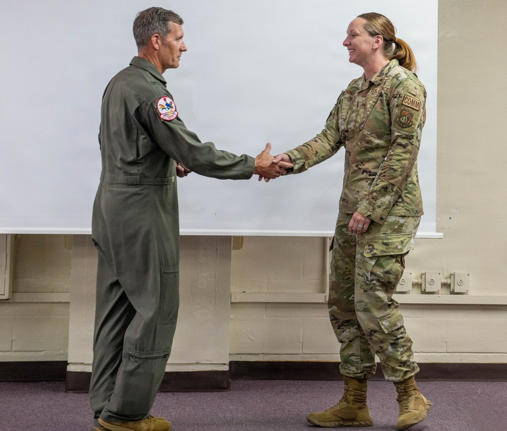 Two U.S. military personnel are shaking hands in what appears to be an indoor briefing or ceremony room. The individual on the left wears an olive green flight suit with a squadron patch on the sleeve, while the individual on the right wears U.S. Air Force OCP (Operational Camouflage Pattern) uniform with 'COMM' and 'U.S. AIR FORCE' tape visible. The interaction suggests a formal recognition, award, or greeting ceremony.