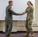 Two U.S. military personnel are shaking hands in what appears to be an indoor briefing or ceremony room. The individual on the left wears an olive green flight suit with a squadron patch on the sleeve, while the individual on the right wears U.S. Air Force OCP (Operational Camouflage Pattern) uniform with 'COMM' and 'U.S. AIR FORCE' tape visible. The interaction suggests a formal recognition, award, or greeting ceremony.
