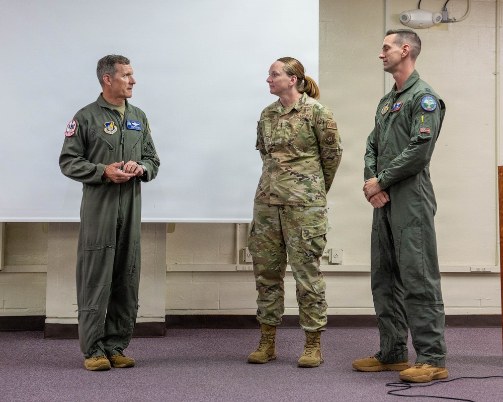 Three U.S. Air Force personnel are standing in what appears to be a briefing or classroom setting. Two individuals are wearing olive green flight suits with unit patches, while one is wearing the standard OCP (Operational Camouflage Pattern) uniform. The person on the left appears to be speaking or addressing the other two.