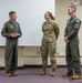 Three U.S. Air Force personnel are standing in what appears to be a briefing or classroom setting. Two individuals are wearing olive green flight suits with unit patches, while one is wearing the standard OCP (Operational Camouflage Pattern) uniform. The person on the left appears to be speaking or addressing the other two.