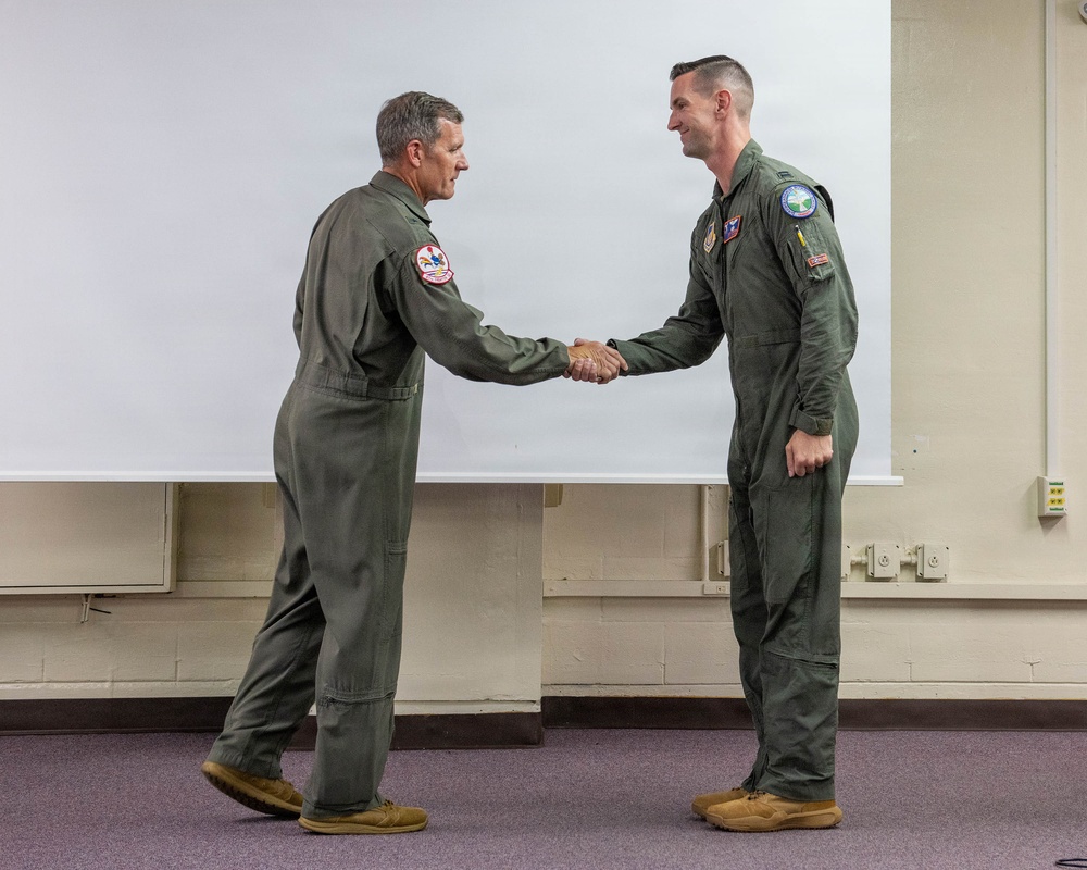 Two military personnel in olive green flight suits are shaking hands in what appears to be a briefing room or classroom setting. Both individuals wear unit patches on their left shoulders, suggesting a formal recognition or ceremonial exchange. The setting and attire are consistent with a U.S. Air Force or similar military aviation unit.