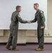 Two military personnel in olive green flight suits are shaking hands in what appears to be a briefing room or classroom setting. Both individuals wear unit patches on their left shoulders, suggesting a formal recognition or ceremonial exchange. The setting and attire are consistent with a U.S. Air Force or similar military aviation unit.