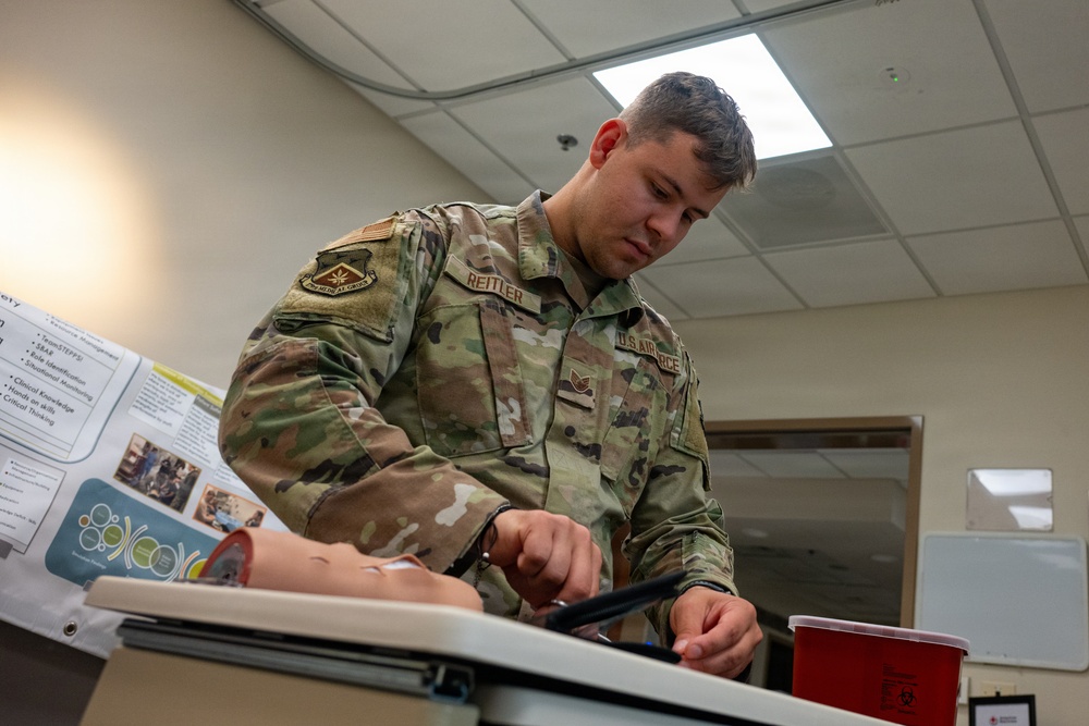 A U.S. Air Force service member named Reitler is practicing a medical procedure on a training mannequin arm, likely an IV insertion or blood draw simulation. The setting appears to be a medical training classroom with instructional posters visible in the background. The soldier is wearing OCP (Operational Camouflage Pattern) uniform with U.S. Air Force insignia.