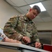 A U.S. Air Force service member named Reitler is practicing a medical procedure on a training mannequin arm, likely an IV insertion or blood draw simulation. The setting appears to be a medical training classroom with instructional posters visible in the background. The soldier is wearing OCP (Operational Camouflage Pattern) uniform with U.S. Air Force insignia.