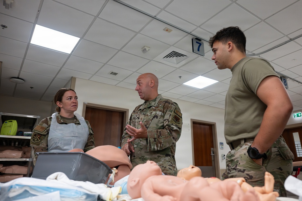 Three military personnel in U.S. Army/Air Force uniforms are gathered around a medical simulation table in what appears to be a medical training facility. A senior uniformed officer in multicam/OCP pattern is speaking and gesturing to the other two personnel. The table contains medical training mannequins including an adult torso and an infant simulator, indicating obstetric or pediatric emergency care training.