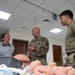Three military personnel in U.S. Army/Air Force uniforms are gathered around a medical simulation table in what appears to be a medical training facility. A senior uniformed officer in multicam/OCP pattern is speaking and gesturing to the other two personnel. The table contains medical training mannequins including an adult torso and an infant simulator, indicating obstetric or pediatric emergency care training.