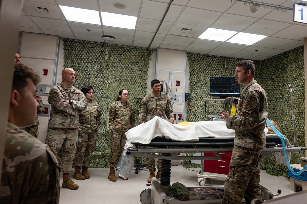A group of military personnel in OCP (Operational Camouflage Pattern) uniforms are gathered in what appears to be a medical training or simulation room. One individual on the right appears to be briefing or instructing the group near a patient gurney covered with a white sheet. The room is decorated with camouflage netting on the walls and contains medical monitoring equipment.