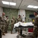 A group of military personnel in OCP (Operational Camouflage Pattern) uniforms are gathered in what appears to be a medical training or simulation room. One individual on the right appears to be briefing or instructing the group near a patient gurney covered with a white sheet. The room is decorated with camouflage netting on the walls and contains medical monitoring equipment.