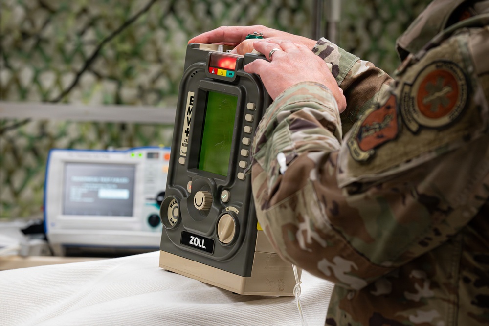 A military personnel member in MultiCam/OCP camouflage uniform is operating a ZOLL medical device, likely a defibrillator or patient monitor, on a white surface. The setting appears to be a field medical station indicated by camouflage netting in the background. A secondary medical monitor is visible in the background, suggesting a medical training or operational environment.