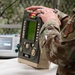 A military personnel member in MultiCam/OCP camouflage uniform is operating a ZOLL medical device, likely a defibrillator or patient monitor, on a white surface. The setting appears to be a field medical station indicated by camouflage netting in the background. A secondary medical monitor is visible in the background, suggesting a medical training or operational environment.