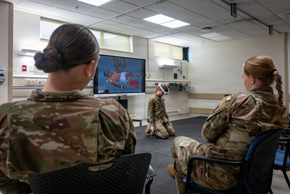 Three military personnel in camouflage uniforms are engaged in a virtual reality medical training exercise. One soldier kneels on the floor wearing a VR headset and holding controllers, while two others observe from chairs. A monitor displays the VR simulation showing a casualty scenario, branded with 'SimX' software.