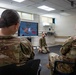 Three military personnel in camouflage uniforms are engaged in a virtual reality medical training exercise. One soldier kneels on the floor wearing a VR headset and holding controllers, while two others observe from chairs. A monitor displays the VR simulation showing a casualty scenario, branded with 'SimX' software.