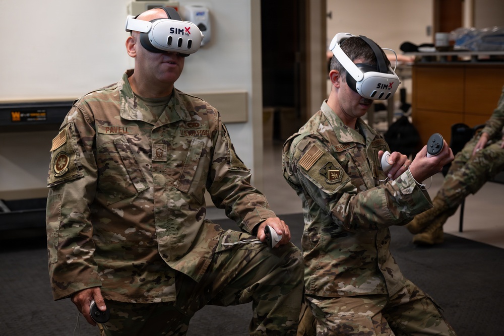 Two U.S. Air Force personnel in OCP (Operational Camouflage Pattern) uniforms are using SimX VR headsets and hand controllers, likely engaged in a virtual reality training simulation. The left individual's name tape reads 'PAVELL' and both wear U.S. Air Force branch tapes. They appear to be in a training facility or classroom environment.