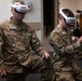 Two U.S. Air Force personnel in OCP (Operational Camouflage Pattern) uniforms are using SimX VR headsets and hand controllers, likely engaged in a virtual reality training simulation. The left individual's name tape reads 'PAVELL' and both wear U.S. Air Force branch tapes. They appear to be in a training facility or classroom environment.