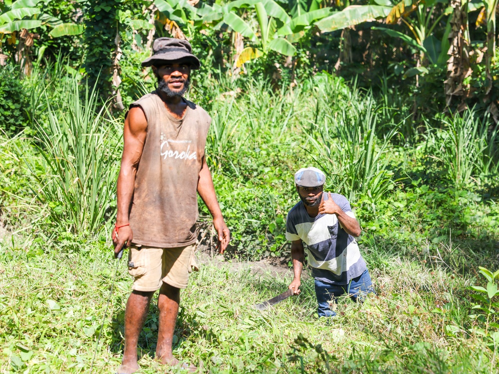 Two men are working in a lush, tropical agricultural area surrounded by dense green vegetation including banana trees. The man on the left is standing upright wearing a sleeveless brown shirt reading 'Goroka' and a bucket hat, while the man on the right is crouching and appears to be holding a machete or cutting tool. Both men appear to be engaged in land clearing or farming work.