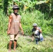 Two men are working in a lush, tropical agricultural area surrounded by dense green vegetation including banana trees. The man on the left is standing upright wearing a sleeveless brown shirt reading 'Goroka' and a bucket hat, while the man on the right is crouching and appears to be holding a machete or cutting tool. Both men appear to be engaged in land clearing or farming work.