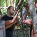 A group of people are gathered in a tropical forest setting, working together around a tree or large branch. The central figure, a muscular man in a black t-shirt with facial markings/paint, appears to be engaged in a hands-on activity with two other individuals who also have body markings and dirt on their skin. The setting and body decorations suggest a cultural immersion or survival/adventure experience.