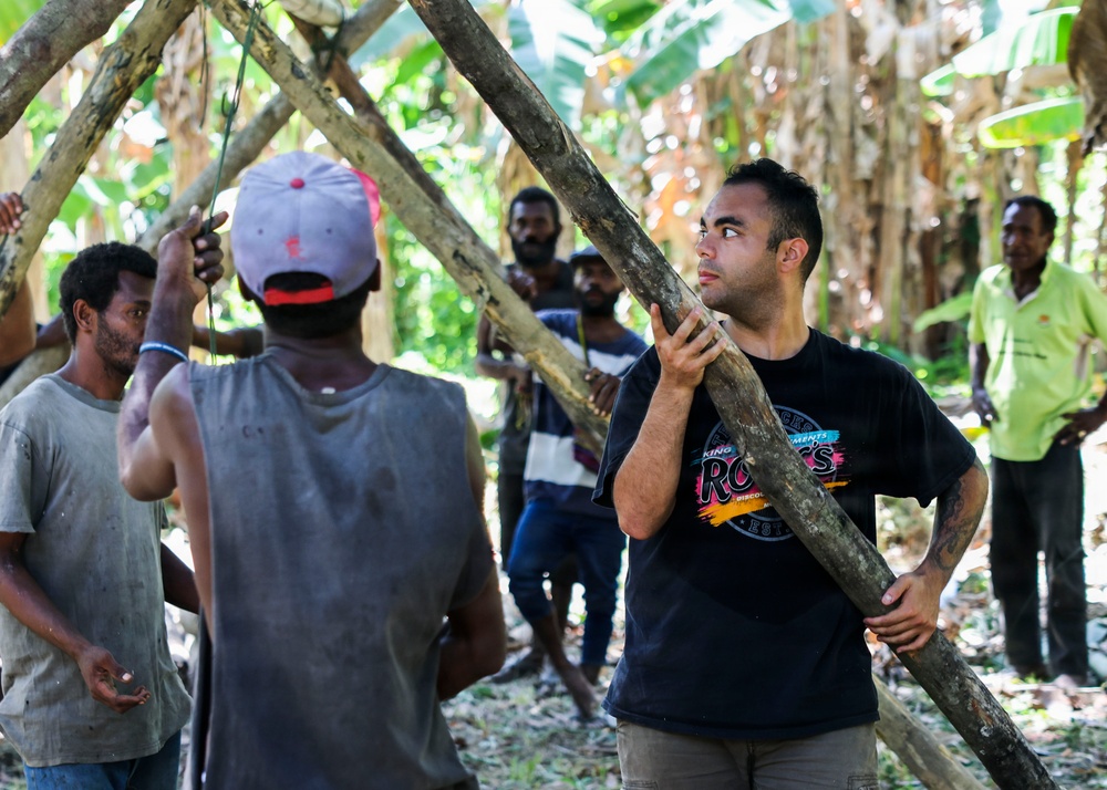 A group of men are working together in a tropical forest setting, constructing a structure using wooden poles and branches. One man wearing a black t-shirt and a tattoo on his arm is holding a large wooden pole, appearing to coordinate with others. The setting appears to be in a tropical region, likely in the Pacific Islands, based on the vegetation and the appearance of the participants.