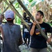 A group of men are working together in a tropical forest setting, constructing a structure using wooden poles and branches. One man wearing a black t-shirt and a tattoo on his arm is holding a large wooden pole, appearing to coordinate with others. The setting appears to be in a tropical region, likely in the Pacific Islands, based on the vegetation and the appearance of the participants.