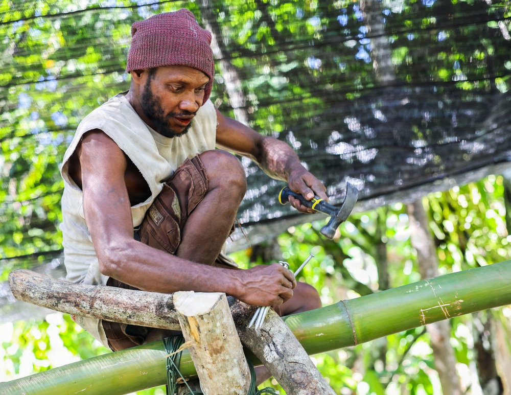 A man wearing a dark red knit cap and sleeveless shirt is seated on bamboo poles, actively working on a construction project. He is holding a hammer in one hand and a nail in the other, appearing to fasten bamboo or wooden structural elements together. The setting is outdoors in a tropical or subtropical environment with lush green vegetation visible in the background.