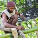 A man wearing a dark red knit cap and sleeveless shirt is seated on bamboo poles, actively working on a construction project. He is holding a hammer in one hand and a nail in the other, appearing to fasten bamboo or wooden structural elements together. The setting is outdoors in a tropical or subtropical environment with lush green vegetation visible in the background.