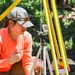 A woman wearing an orange long-sleeve shirt and a gray cap with sunglasses is crouching outdoors, closely examining a surveying or measuring instrument mounted on a silver tripod. Yellow survey poles or range rods are visible in the foreground and background, suggesting a field survey or environmental measurement activity. The setting appears to be a vegetated outdoor area with natural lighting.