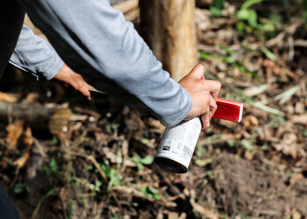 A person wearing a gray long-sleeve shirt is holding a cylindrical can or aerosol container with a red cap/nozzle in an outdoor wooded setting. The background shows forest floor debris including twigs, leaves, and soil. The focus is on the hands and the object being held.