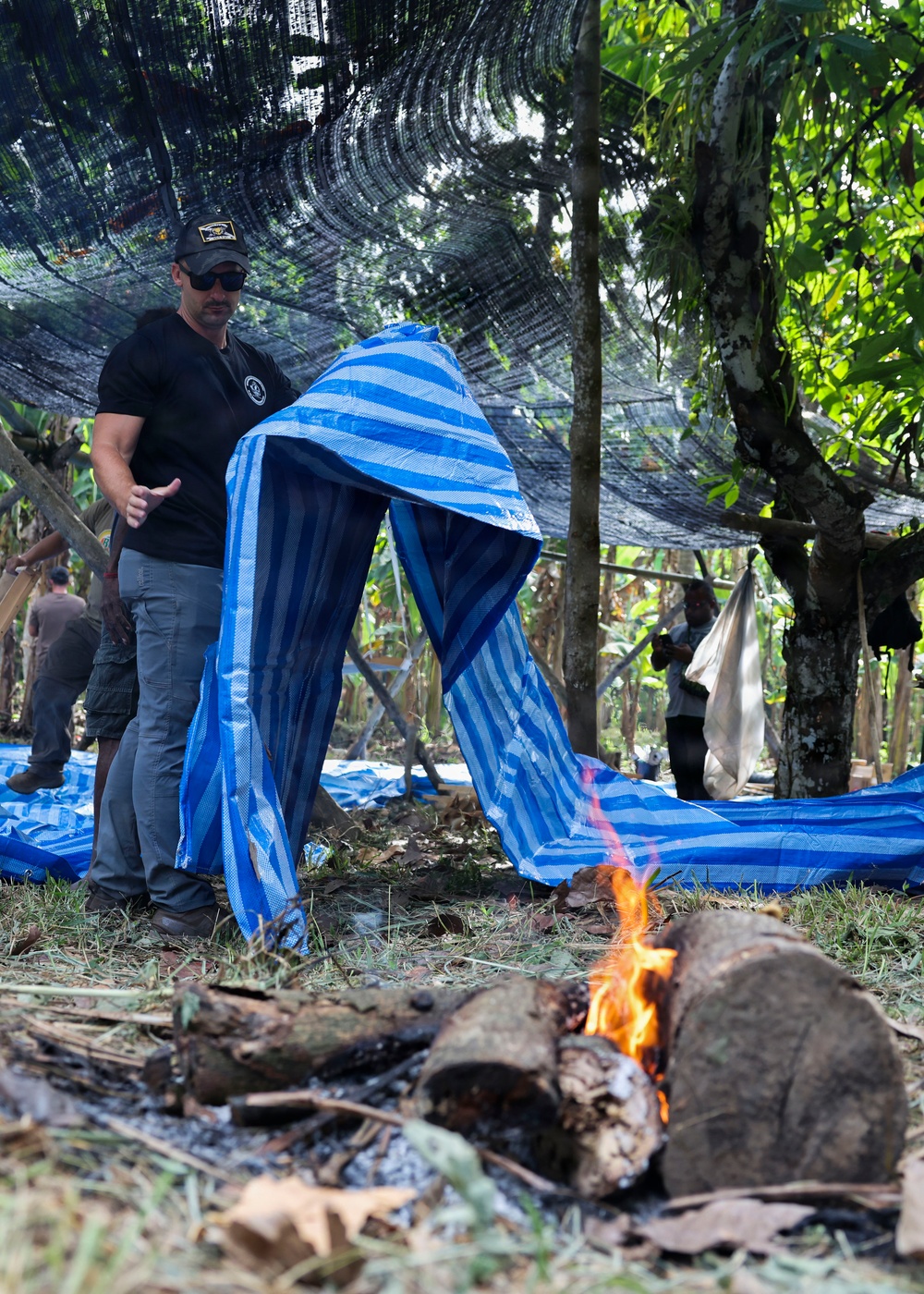 A man wearing a black t-shirt with a logo patch, sunglasses, and a cap is handling a large blue and white striped tarp or woven bag in an outdoor jungle/forest setting. A small fire burns on logs in the foreground, while other individuals are visible in the background among trees covered by a dark camouflage netting. The scene appears to involve some form of field operation or camp activity.