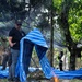 A man wearing a black t-shirt with a logo patch, sunglasses, and a cap is handling a large blue and white striped tarp or woven bag in an outdoor jungle/forest setting. A small fire burns on logs in the foreground, while other individuals are visible in the background among trees covered by a dark camouflage netting. The scene appears to involve some form of field operation or camp activity.