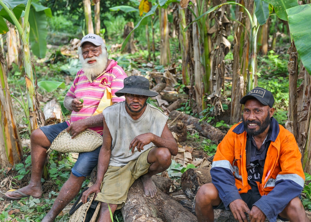 Three men are seated outdoors in a lush tropical setting surrounded by banana trees and cut logs. The group appears relaxed and at ease, with one man smiling broadly at the camera. The setting suggests a rural or village environment, likely in the Pacific Islands or a similar tropical region.