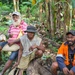 Three men are seated outdoors in a lush tropical setting surrounded by banana trees and cut logs. The group appears relaxed and at ease, with one man smiling broadly at the camera. The setting suggests a rural or village environment, likely in the Pacific Islands or a similar tropical region.