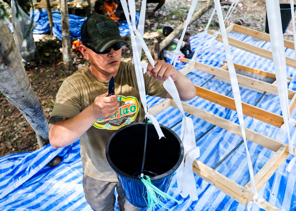 A man wearing a green cap, sunglasses, and an olive t-shirt with a 'Berlao' logo is using a knife to cut or work with white plastic straps attached to a black bucket. He is working within a wooden frame structure covered with blue tarps, likely in an outdoor construction or assembly setting. Additional people and materials are visible in the background.