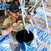 A man wearing a green cap, sunglasses, and an olive t-shirt with a 'Berlao' logo is using a knife to cut or work with white plastic straps attached to a black bucket. He is working within a wooden frame structure covered with blue tarps, likely in an outdoor construction or assembly setting. Additional people and materials are visible in the background.